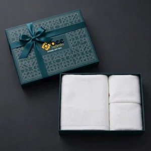 Dark teal patterned gift box lid tied with a satin ribbon bow beside an open box of neatly folded white towels on a dark charcoal background, showing custom apparel gift boxes