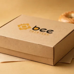 Hero shot of a flat kraft bagel box with the Bee Printers logo printed on the closed lid, warm golden-beige studio backdrop