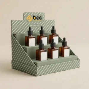 Sage green tiered counter display with white diagonal stripes holding six square amber dropper bottles with white labels on a beige background with the Bee Printers logo on the header, showing custom beard oil display boxes