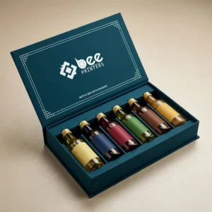 Open dark teal hinged box with Bee Printers logo inside the lid holding six small bottles with colourful labels separated in divider slots on a beige background, showing custom bottle boxes with dividers