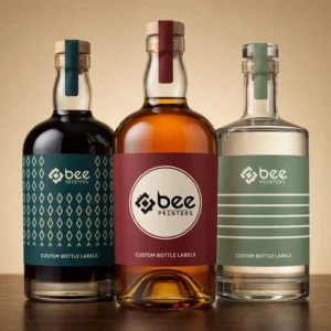 Three glass spirit bottles with dark, amber and clear liquids wearing teal patterned, burgundy circular and sage striped labels on a warm beige background with the Bee Printers logo, showing custom bottle labels
