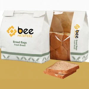 Two white paper bags with green wheat trim, one closed and one with a clear window showing a loaf, with sliced bread in front on a mustard-yellow surface, showing custom bread bags by Bee Printers.