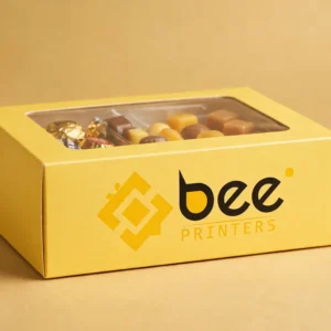 Custom candy boxes in bright printed board with a clear window lid and the Bee Printers logo printed on the front, closed hero box on a warm golden-beige studio backdrop