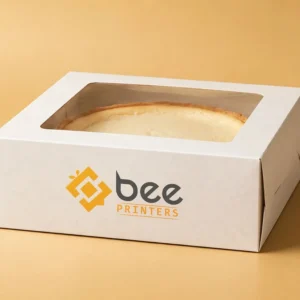 Custom cheesecake boxes in white board with a clear window lid and the Bee Printers logo printed on the side, closed hero box on a warm golden-beige studio backdrop