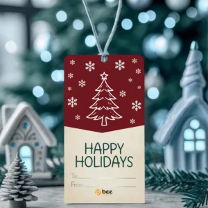 A red and cream rectangular tag with a white Christmas tree outline, snowflakes and Happy Holidays To and From lines hanging on white twine before a blurred festive background with miniature houses, showing custom christmas gift hang tags with a small Bee Printers logo at the base