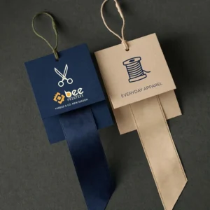 Two square folded tags in navy blue and kraft beige with scissors and thread spool icons, satin ribbons and cord loops on a dark grey surface, showing custom clothing hang tags with the Bee Printers logo on the navy tag