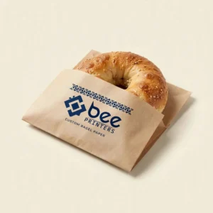 A kraft paper bagel sleeve with a navy Bee Printers logo holding a sesame bagel on a cream background, showing custom custom bagel paper