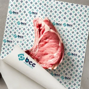 White butcher paper with a teal and navy diamond pattern and repeating teal-mark Bee Printers logos, one corner rolled up, holding a large raw steak on a grey surface, showing custom custom butcher paper