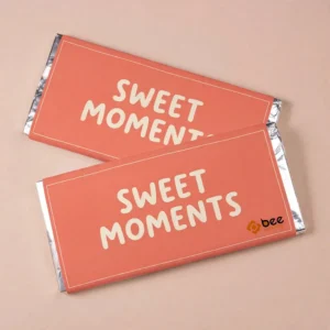Two coral-orange wrapped chocolate bars with cream SWEET MOMENTS lettering and silver foil ends on a pale beige background, showing custom custom chocolate wrapping paper