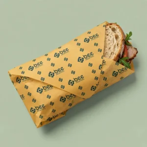 A sandwich wrapped in mustard yellow deli paper with a repeating dark teal Bee Printers logo pattern on a pale sage background, showing custom custom deli paper