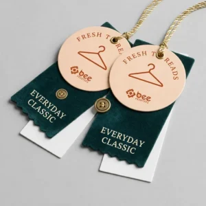 Two round peach tags with a hanger icon, Fresh Threads text and gold cord resting on dark green Everyday Classic fabric swatches with brass buttons on a light grey surface, showing custom die cut hang tags with a burnt orange Bee Printers logo