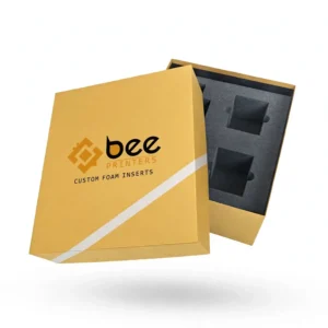 A mustard yellow box lid with a white diagonal stripe and the Bee Printers logo on its front leaning against an open box revealing a black foam insert with square cutouts on a white background, showing custom foam inserts for boxes
