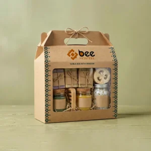 A kraft-brown custom gable boxes with window design tied with twine, its window showing wrapped soaps, small jars and a loofah, with a teal diamond border and the Bee Printers logo on a sage-green background.