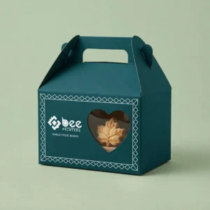 Closed dark teal gable box with a white diamond border and a heart-shaped window showing a leaf-shaped biscuit on a pale green background, showing custom gable food boxes.