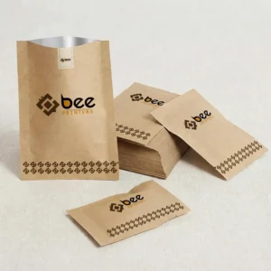 Kraft heat seal paper pouches in upright, stacked and flat sealed sachet form with a dark brown geometric border pattern and Bee Printers logo on a light fabric background, showing custom heat seal papers.