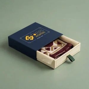 A navy blue drawer-style apparel box partly open with the Bee Printers logo on top of the sleeve, revealing a cream tray holding a folded burgundy and cream patterned garment, on a solid sage green background, showing custom luxury apparel boxes.