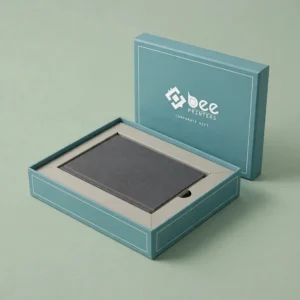 An open teal two-piece business gift box with its lid standing behind it holding a dark grey flat notebook inside, with a white Bee Printers logo on the lid, against a plain sage green background, showing custom luxury business gift boxes.
