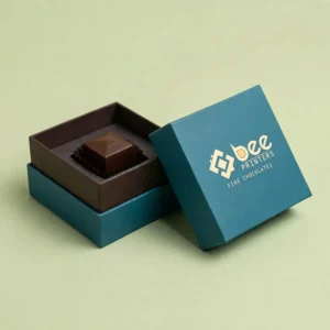An open teal and brown chocolate box with its lid leaning beside it holding a single square chocolate inside, with a cream Bee Printers logo on the lid, against a plain pale green background, showing custom luxury chocolate boxes.