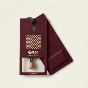 Two burgundy rectangular tags with a gold lattice pattern, cream Bee Printers logo, Premium Quality text and a rectangular window cut-out with a rose gold cord lock on a cream background, showing custom luxury hang tags