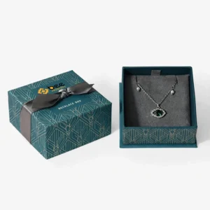 A teal geometric-patterned necklace box with a black ribbon bow beside its open tray holding a silver necklace with a green pendant on a grey cushion on a white background, showing custom luxury necklace boxes