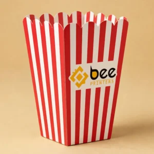 Hero shot of a classic red-and-white striped popcorn box with the Bee Printers logo printed on the front, warm golden-beige studio backdrop