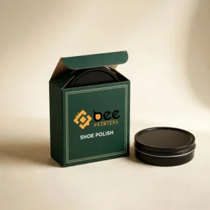 An open dark green box from the custom shoe polish packaging boxes range sits on a beige background with a black shoe polish tin inside and another tin beside it, printed with the Bee Printers logo and SHOE POLISH text.