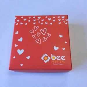 A closed red square box viewed from above on a pale lavender background, decorated with white filled and outlined hearts, with a white and yellow Bee Printers logo and Sweet Treats text at the lower right of the lid, showing custom valentine candy boxes.
