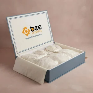 Open dusty blue custom wedding dress storage boxes with the Bee Printers logo inside the hinged lid, holding a folded ivory lace wedding dress tied with cream ribbons on a dusty pink background.