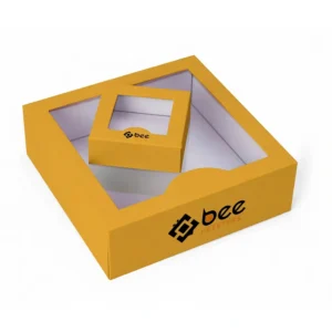 A large mustard-yellow shallow window box with a clear window and white empty interior holding a smaller matching window box on a white background, with the Bee Printers logo in black and orange on the front side and a smaller logo on the small box, showing custom window boxes.